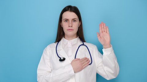 Honest woman doctor, intern giving pledge, raising one arm and hold hand on heart while promise, oath to patient, isolated on blue wall. Covid 19, healthcare workers and preventing virus concept.