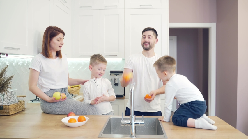 Cheerful young family with small children juggling fruits in kitchen. Two little boys help their parents with cooking. Spend time together at home. Funny dad and mom have fun with kids at home Royalty-Free Stock Footage #1066761796