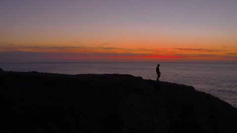 Silhouette of lonely single man walk on edge of cliff, to watch over last sun light at sunset dusk. Epic cinematic drone shot of isolated man immersed in self care. Tranquility in nature, small human