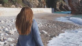 Camera follows beautiful redhead woman in light blue coat on the seaside. Hands-on video. Film grain