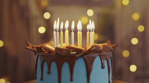 Birthday Cake with Burning Candles, chocolate glaze is Rotating on Stand on Table in House. Bright Dessert for Child's Birthday Party on Background of Bokeh Light Bulbs Garlands. Family. Childhood