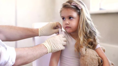 Immunization for children concept. Happy little cute blonde girl holding a toy and getting a flu shot not afraid of the syringe needle. Doctor injecting brave child with Covid-19 vaccine at clinic or