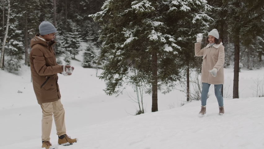 Wide shot of young African-American man playing snowballs with his female mixed-race friend while having walk in forest in winter