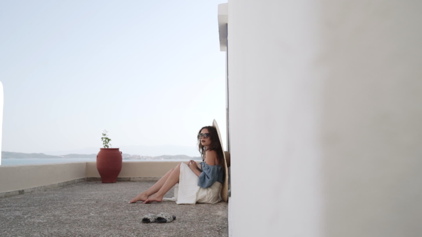Long shot of young woman sitting down on the ground in a light white dress and watching a beautiful sea scenery.