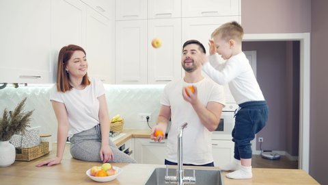 Cheerful young family with small children juggling fruits in kitchen. Two little boys help their parents with cooking. Spend time together at home. Funny dad and mom have fun with kids at home