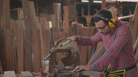 Handsome carpenter man wearing goggles and earmuffs for personal safety using circular saw cut wood for furniture in workplace at factory. Concept of small business and owner startup about woodwork. 