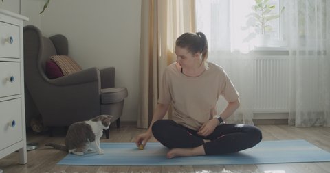 Young woman in sportswear sitting in a lotus position and playing with a cat during workout exercise at home.
