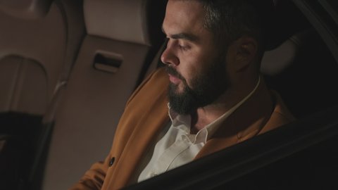 Handheld closeup of mixed race businessman answering phone call, chatting while sitting in backseat of luxury black car at night