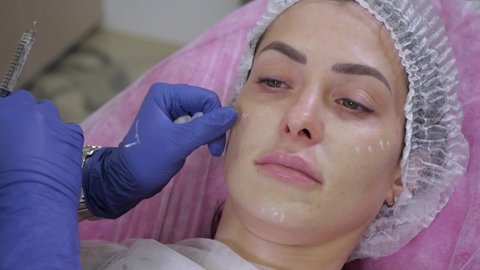 The doctor of the beautician in medical gloves makes a cosmetic procedure to the girl patient injections to rejuvenate the skin in the line of cheeks with a medical syringe in the cosmetics office