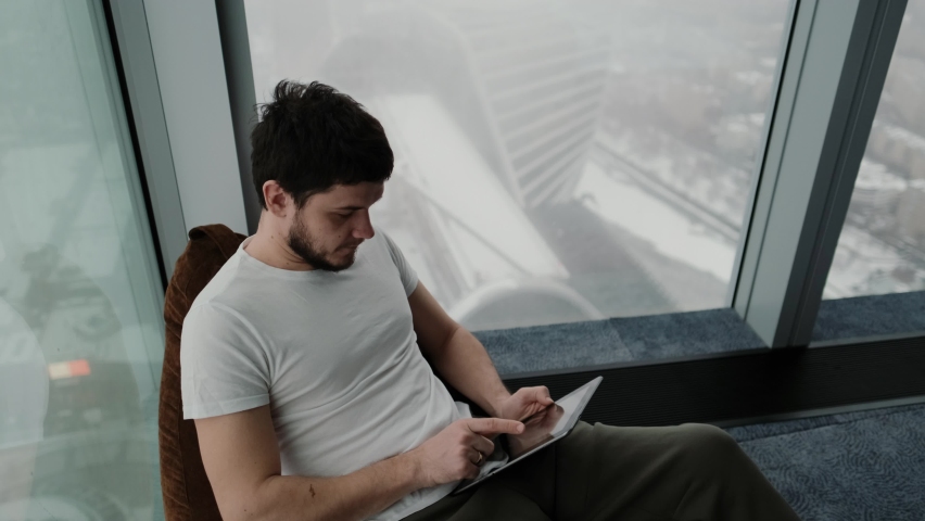 Gorgeous man sitting on pouf using tablet in modern apartment, texting, scrolling through technologies, isolated lifestyle