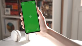 Girl using smartphone with green screen in living room watching movie, video content. Tracking markers. Smart phone in a vertical position. Indoors. Closeup. Blurred room on the background