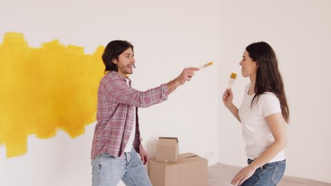 Funny repair. Young married man and woman painting walls at home, trying to paint each other, laughing and having fun, slow motion