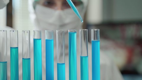 Female laboratory researcher in protective suit sitting on background of monitor with moving microbe video and dropping blue reagent in sterile test-tubes. Professional virologist holding important