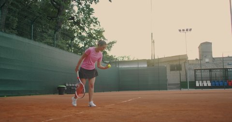 The girl took her stance before serving. Smash, after bouncing, she hits the tennis ball near the ground with a right hand, A Clay court with a green fence. Wide angle, through the mesh.