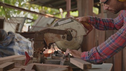Close up of handsome carpenter man wearing goggles and earmuffs for personal safety using circular saw cut wood for furniture in workplace. Concept of small business and owner startup about woodwork.