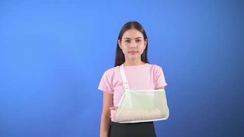Portrait of young woman with an injured arm in a sling over blue background in studio, insurance and healthcare concept