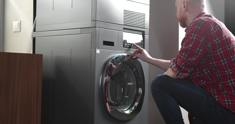 Side view of young man loading dirty clothes into washing machine. Handsome guy doing laundry and starting washing machine in bathroom or launderette