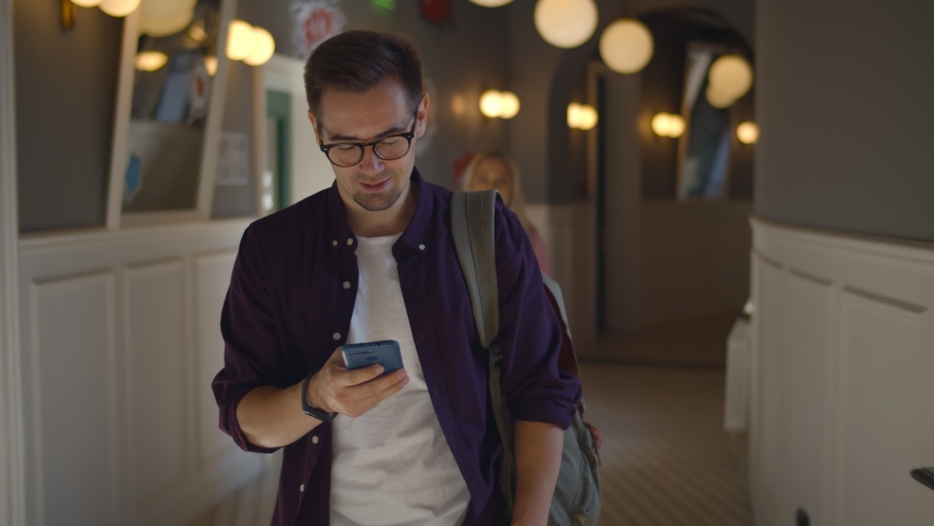 Cheerful male student surfing internet on smartphone walking in university hall. Portrait of college hipster guy with backpack chatting on mobile phone walking indoors