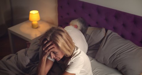 Senior woman sitting in bed and feeling stressed with husband sleeping on background. Portrait of aged worried woman having insomnia and anxiety while husband sleeping