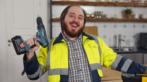 Portrait of funny male worker in uniform with screwdriver in kitchen. Overweight handyman holding electric drill and smiling at camera standing in modern kitchen with toolbox