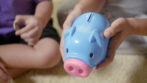 Happy Asian kid boy preschool and mother putting pin money coins into piglet. Little child putting coin into piggy bank for saving with pile of coins at home, Investment education concept, Slow motion