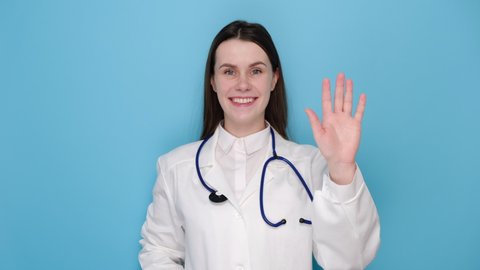 Portrait of friendly young doctor woman wearing medical uniform waving saying hello happy and smiling, welcome gesture, isolated on blue studio background. Covid 19, virus, health and medicine concept