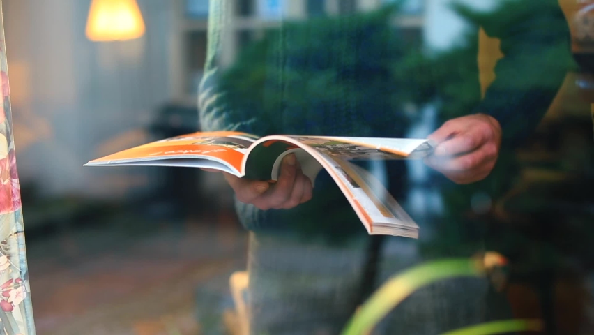 Man leafs through magazine near window in self-isolation house. He turns page with hand. Use printed publications on paper. Camera movement through glass, glare, reflections. Power of education  Royalty-Free Stock Footage #1066827493
