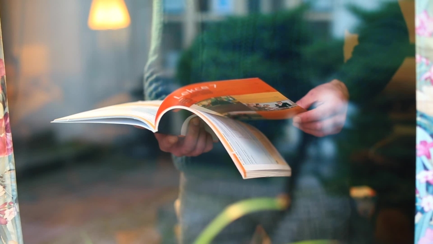 Man leafs through magazine near window in self-isolation house. He turns page with hand. Use printed publications on paper. Camera movement through glass, glare, reflections. | Shutterstock HD Video #1066827493
