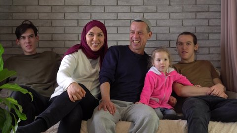 A happy middle eastern Muslim family sits together on the sofa and laughs. Ramadan holiday. Woman in hijab and man with children. Mother father with teen sons and little daughter