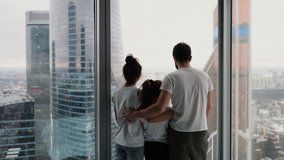 Young family with daughter spending time together in modern apartment with large windows overlooking the city from a high floor.