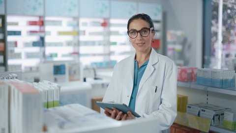 Pharmacy Drugstore: Beautiful Caucasian Pharmacist Uses Digital Tablet Computer, Checks Inventory of Medicine, Drugs, Vitamins, Health Care Products on a Shelf. Professional Pharmacist in Pharma Store
