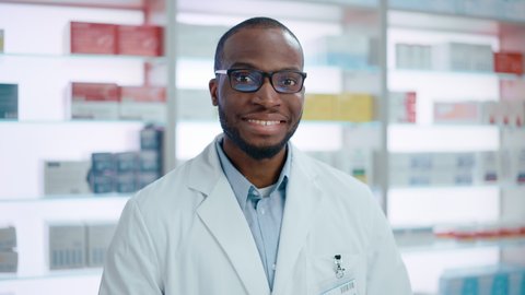 Pharmacy Checkout Counter: Portrait of Handsome Black Male Pharmacist, Slowly Rising His Eyes, Looks at Camera Smiling Charmingly. Drugstore with Shelves Health Care Products, Medicine. Slow Motion
