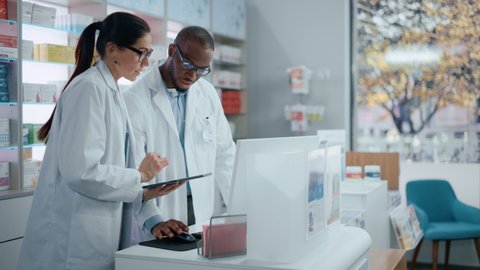 Pharmacy Behind the Checkout Counter: Black Male and Caucasian Female Pharmacists Talk about Medicine, Use Digital Tablet Computer. Medical Professionals in Drugstore with Health Care Products