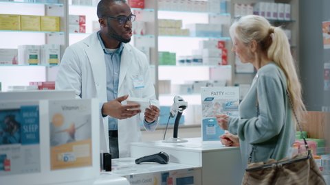 Pharmacy Drugstore Checkout Counter: Black Male Pharmacist Explains Use and Manual for Prescription Medicine Beautiful, Senior Female Customer Paying Using Contactless Credit Card to Terminal