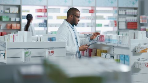 Pharmacy: Portrait of Professional Black Pharmacist Uses Digital Tablet Computer, Checks Inventory of Medicine, Drugs, Vitamins, Health Care Products. Druggist in Drugstore Store. Focus on Hands