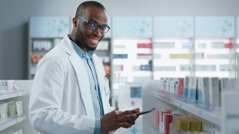 Pharmacy: Portrait of Professional Black Pharmacist Uses Digital Tablet Computer, Checks Inventory of Medicine, Looks at Camera and Smiles Charmingly. Drugstore Store With Health Care Products