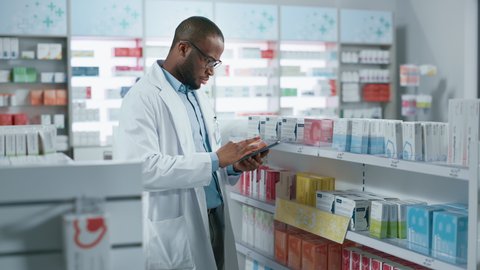 Pharmacy: Portrait of Professional Black Pharmacist Uses Digital Tablet Computer, Checks Inventory of Medicine, Drugs, Vitamins on a Shelf. Drugstore Store Arranging Health Care Products. Zoom in Shot