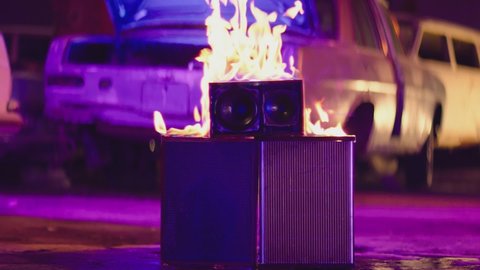 Scene of burning speaker in abandoned garage. Purple and blue lights. Speaker is burning on the concrete floor in the street. Fire burning in slow motion. Vintage and retro style.