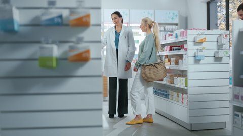 Pharmacy: Professional Pharmacist Helping Beautiful Senior Female Customer with Medicine Recommendation, Advice, Talking. Drugstore with Full of Drugs Packages, Pills Bottles, Health Care Products