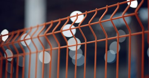 Modern orange fence made of metal mesh against the background of bright lights in the blur in the dark