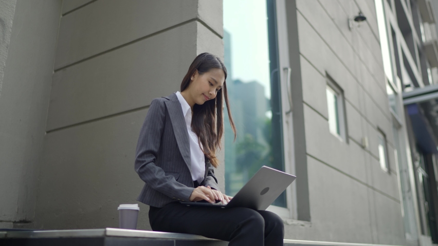 Video Slow-motion. A beautiful white-skinned Asian woman in a black suit sitting in front of the office building. Using the laptop on the side of the road in the city. Royalty-Free Stock Footage #1066848499