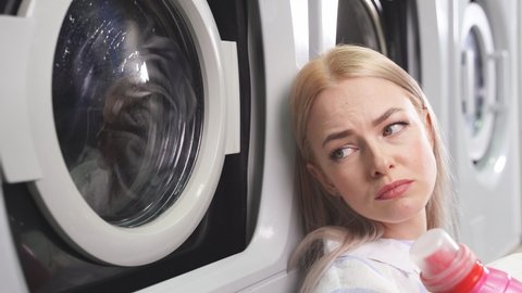 Close-up of a young woman with a sad expression sitting by the washing machine in a public laundry.