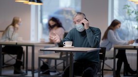 Senior man in coronavirus face mask talking on the phone sitting in restaurant with blurred people at background. Portrait of male Caucasian retiree dining in cafe on Covid-19 pandemic. New normal. - Powered by Shutterstock - Get 15% off with code: PIKWIZARD15