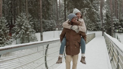 Lockdown of young African-American man carrying on his back his attractive girlfriend while walking in woods in winter and enjoying view Video stock