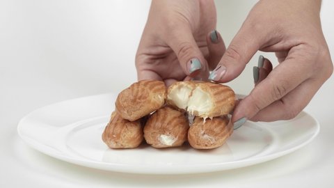 Women's hands break the eclair into two halves. Beautiful female hands share the eclair, showing the filling Close-up. Eclairs on a white round plate on a white background.