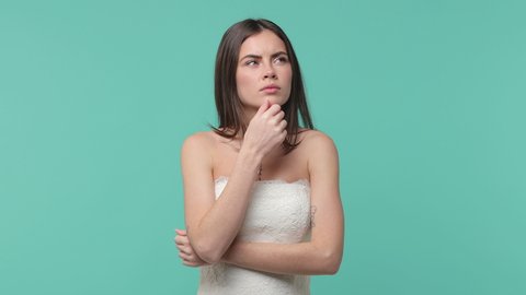 Puzzled pensive bride young brunette woman 20s years old in beautiful white wedding dress posing isolated on blue turquoise background studio. Wedding concept. Looking aside put hand prop up on chin