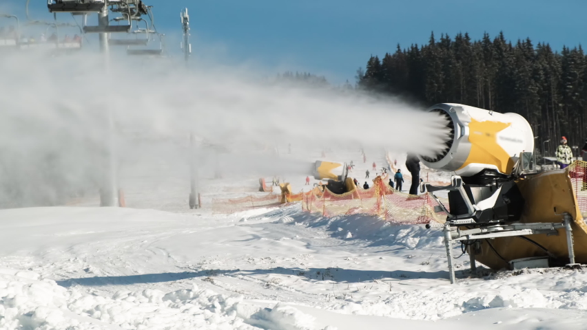 Snow making system. Snow cannon makes artificial snow of water on sunny day in ski resort. On backdrop skiers slide down snow-covered white slope of mountain, ski lift working, winter forest, blue sky