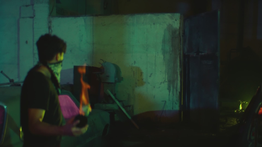 Young male protester covered his face with black bandanna mask   throwing bottle of Molotov cocktail in concrete wall on abandoned building at night. House burning down on fire. Slow motion shot.  