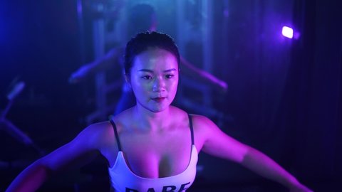 Asian woman sitting doing lateral raises from above. Smoky dark and moody gym with young girl alone doing strength training back exercises with dumbbell. Colored atmospheric fitness center.