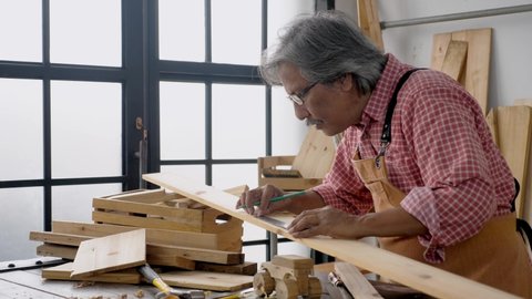 Side view, medium shot portrait of dedicated Asian senior carpenter wears lumberjack shirt and apron, measuring and marking on plywood slat with pencil, making furniture at workshop.